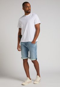 T-shirt blanc à manches courtes, shorts en denim bleu clair avec revers, et baskets beiges avec lacets blancs et accents rouges.