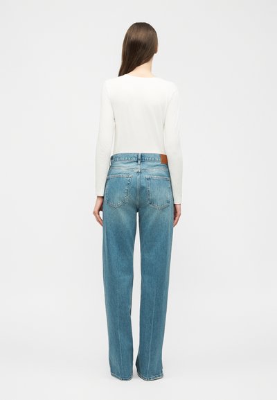 Woman with long brown hair wearing a white long-sleeve top and high-waisted straight blue jeans, standing facing away on a white background.