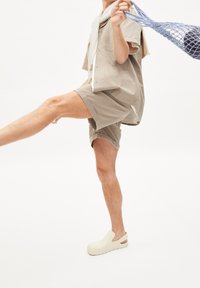 Camicia corta in lino beige con tasca, abbinata a pantaloni corti coordinati e zoccoli color crema con ritagli laterali. Modella che tiene una borsa a rete blu.