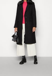 Abrigo de lana negro con cuello de piel sintética, diseño de botonadura simple, combinado con un suéter rosa, pantalones de pierna ancha blancos y un pequeño bolso negro.