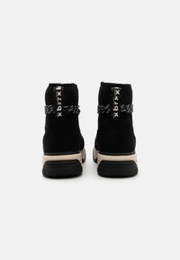 Bottines en suède noir avec une semelle en caoutchouc texturée, présentant un détail décoratif en corde noire et blanche et une sangle de marque au talon.