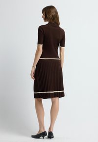 Brun strikket kjole med tætsiddende overdel og plisseret nederdel, fremhævet med cremefarvede striber ved ærmerne og kanten. Sorte højhælede sko.