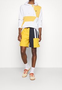 Felpa bianca e gialla con cappuccio e motivi geometrici, abbinata a pantaloni corti gialli e blu navy con tasca. Calzature casual completano l'outfit.
