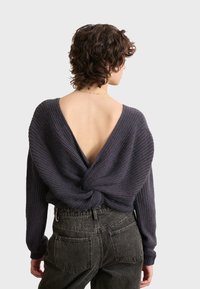 Dunkelgrauer Strickpullover mit tiefem V-Ausschnitt hinten, an der Taille gerafft. Lange Ärmel aus strukturiertem Stoff, kombiniert mit hochgeschnittenen Jeans.