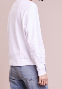 Osoba oblečená v jednoduché bílé mikině s dlouhým rukávem a světle modrých džínách, stojící proti neutrálnímu béžovému pozadí.