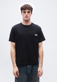 Νέος άνδρας με κοντά, σγουρά μαλλιά, που φοράει μαύρο τζινάκι Calvin Klein με τσέπη και μπλε τζιν, μπροστά σε απλό λευκό φόντο.