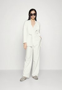 Valkoinen räätälöity puku, jossa syvä V-kaula-aukko ja leveälahkeiset housut, yhdistettynä mustiin aurinkolaseihin ja paljettikoristeisiin slip-on-kenkiin. Sileä kangaspinta.