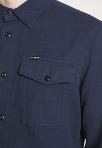 Camicia button-up blu navy realizzata in tessuto testurizzato. Presenta una tasca sul petto e un piccolo etichetta del marchio. I bottoni neri completano il design.