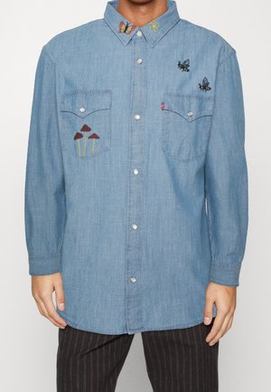 Lyseblå denimskjorte med forlommer, broderede svampe på venstre lomme, to bier på højre bryst og en sommerfugl på kraven.