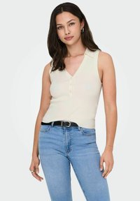 Top senza maniche a costine color crema con scollatura a V e chiusura a bottoni, abbinato a jeans azzurri a vita alta e una cintura nera.
