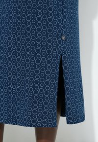 Falda azul marino con patrón circular cosido en blanco, con abertura lateral y pequeña etiqueta de marca negra cerca de la abertura, usada por una persona.