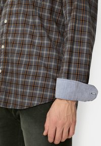 Button-up shirt featuring a dark gray plaid pattern with orange and white lines. Blue chambray cuff, contrasting with the main fabric's texture.