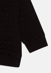 Sivi rukav sweatshirt-a od teksturirane tkanine, s rebrastim manžetama i suptilnim izvezenim "cK" logotipom u usklađenoj niti. Mekan, kutni dizajn.