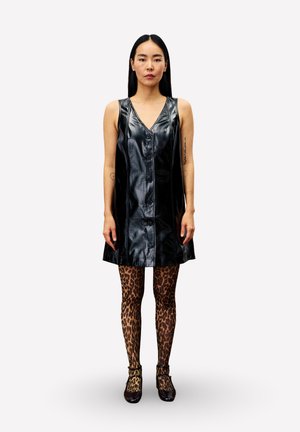 Femme se tenant debout, portant une robe noire en cuir sans manches à boutons et des collants à imprimé léopard avec des chaussures sombres, devant un fond uni.
