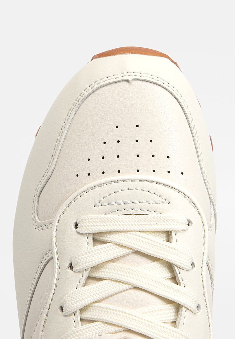 Baskets blanches en cuir avec une finition lisse, des perforations sur le bout, et des lacets crème. Présente un accent de talon marron clair.