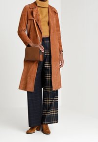 Brun ruskinds trenchcoat over en sennepsfarvet turtleneck, parret med brede plaidbukser og brune ankelstøvler. En matchende taske fuldender looket.
