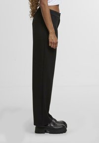 Kalhoty s vysokým pasem v černé barvě se rovnými nohavicemi, jemnými předními sklady a bočními kapsami, doplněné o masivní černé platformové boty.