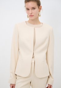 Béžový strukturovaný blazer s dlouhými rukávy, kulatým výstřihem a hladkým povrchem. Obsahuje dvě přední kapsy a skryté zapínání.