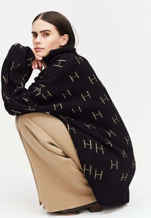 Übergröße schwarzer Pullover mit beigem Logo-Muster, geripptem Kragen und langen Ärmeln. Kombiniert mit weiten beigen Hosen und brauen Schuhen.