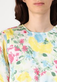 Blouse fleurie en tissu léger et transparent, avec un motif multicolore de fleurs roses, jaunes, bleues et vertes sur fond blanc.