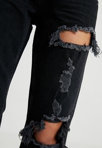 Jeans in denim nero con ginocchia strappate, caratterizzati da bordi sfilacciati e un aspetto usurato. Aderiscono perfettamente alla gamba con un design elegante.