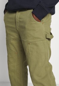 Persoon in olijfgroene cargobroek met zijzakken en een marineblauwe sweatshirt, handen in de voorzakken gestoken.