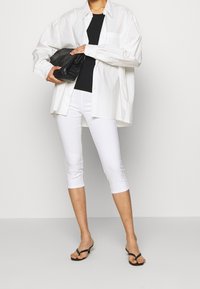 Bílá oversized košile přehozená přes černý přiléhavý top, doplněná bílými capri kalhotami a černými žabkami. Modelka drží černou kabelku.
