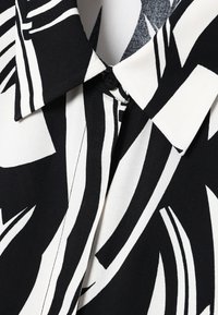 Chemise à motifs noir et blanc avec un col et des boutons, en tissu lisse avec un motif abstrait rappelant des feuilles.