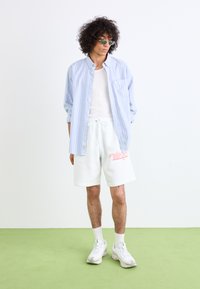 Hvide shorts med pink logo, lyseblå stribet skjorte med knapper og hvide sneakers. Model iført solbriller, stående på grøn overflade.