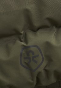 Texturerad olivgrön tyg med subtila veck, med en mörk logga i form av ett sköld. Materialet verkar slitstarkt med en slät yta.