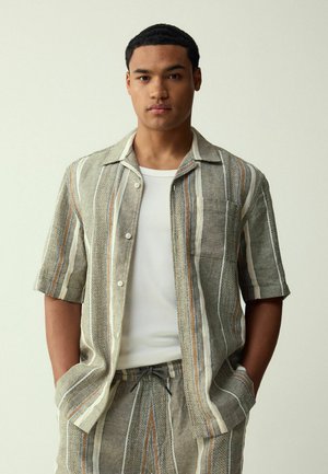 Grau gestreiftes Kurzarmhemd aus Leinen mit mehrfarbigen Akzenten und einer Brusttasche. Kombiniert mit hellgrauen Hose mit Kordelzug.
