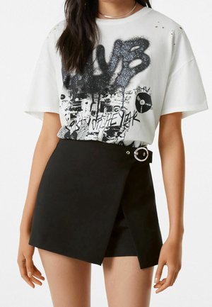 Hvid grafisk T-shirt med sort og sølvfarvet tryk, stylet med en sort asymmetrisk mininederdel med en rund spænde. Blødt stof, afslappet pasform.