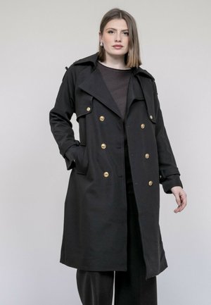 Femme aux cheveux raides mi-longs portant un manteau noir croisé à boutons dorés et un pantalon noir, debout devant un fond uni.