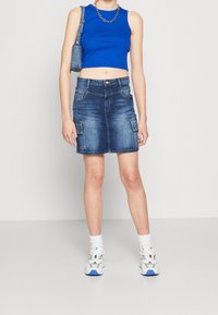 Osoba oblečená v modrém bezrukávovém topu, džínové mini sukni s kapsami, bílých podkolenkách, bílých teniskách a nese malou modrou kabelku přes rameno.