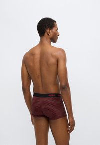 Boxers vermelhos com padrão, com um cós elástico preto, apresentando o nome da marca "HUGO" em destaque. O material parece suave.