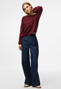 Burgundy neulepusero, jossa on rento malli, yhdistettynä korkeavyötäröisiin, leveälahkeisiin tummansinihin farkkuihin. Silkkinen pinta, hillityt ompeluyksityiskohdat.