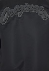 Černá bunda s texturovaným, vystouplým logem "Originals" v kurzívním písmu, které doplňuje hladký povrch látky.