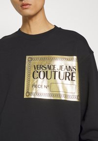 Svart sweatshirt med en guldmetallisk logografik som har texten "VERSACE JEANS COUTURE" och en dekorativ repkant.