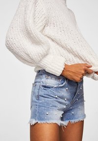 Suéter blanco de punto con mangas abullonadas, combinado con pantalones cortos de mezclilla azules con adornos de perlas y un dobladillo deshilachado.