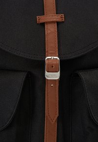 Zwarte stoffen rugzak met een bruine leren riem en een zilverkleurige gesp, voorzien van twee voorzakken en details in de stiksels.