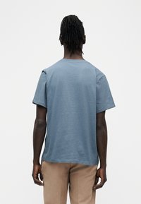 Άντ ρας με κοτσίδες ντρέντλοκς, ντυμένος με μπλε κοντομάνικο μπλουζάκι και μπεζ παντελόνι, στέκεται με την πλάτη προς το μέρος μας μπροστά σε απλό λευκό φόντο.