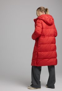 Rode donsjacket met capuchon, voorzien van een gequilt ontwerp en elastische manchetten; gedragen over een grijze broek en casual schoeisel.