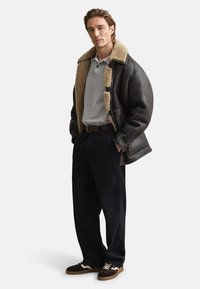 Ruda odinė striukė su kailiu apykaklėje, pilka polo marškinėliai, tamsios kelnės ir rudi sportbačiai su baltais akcentais. Plonas siluetas ir laisvalaikio stilius.