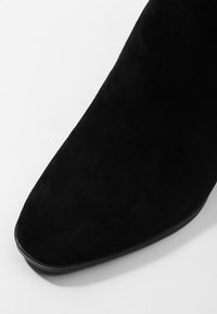 Stivale alla caviglia in suede nero con punta affilata e texture liscia. Presenta una suola bassa e piatta e un design minimalista senza hardware o accenti visibili.