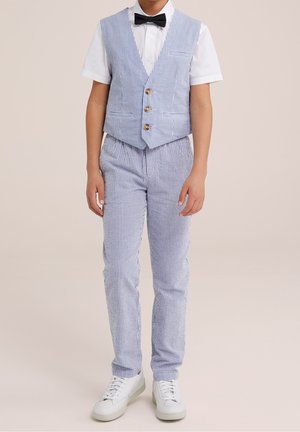 Chaleco a rayas azul y blanco con tres botones, combinado con pantalones a rayas a juego y zapatillas blancas. Camisa blanca de manga corta debajo.