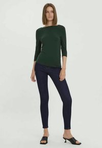 Dunkelgrünes, figurbetontes Oberteil mit drei Viertelärmeln, kombiniert mit dunkelblauen Skinny-Jeans und schwarzen, offenen Mules.