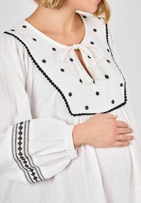 Balta, laisvai krentanti bliuzono forma su juodais gėlėmis išsiuvinėjimu, raktų skylutės atidarymas, surištas balta kutu, ir detali juoda apdaila ant rankovių.