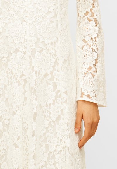 Λευκό φόρεμα από δαντέλα με λουλούδια και μακριά μανίκια, που δείχνει λεπτομερή υφή υφάσματος και ένα χέρι που ακουμπά δίπλα στο σώμα.