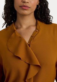 Μπλούζα σε μουσταρδί χρώμα με ριχτό γιακά και κουμπιά κατά μήκος του λαιμού, φτιαγμένη από μαλακό ύφασμα με ελαφριά υφή.