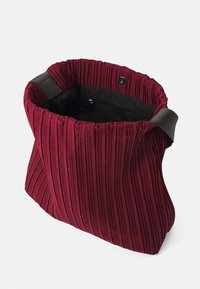 Sac fourre-tout rouge plissé avec un extérieur en tissu doux, ouverture sur le dessus, et un intérieur sombre comprenant une poche zippée et une bandoulière robuste.
